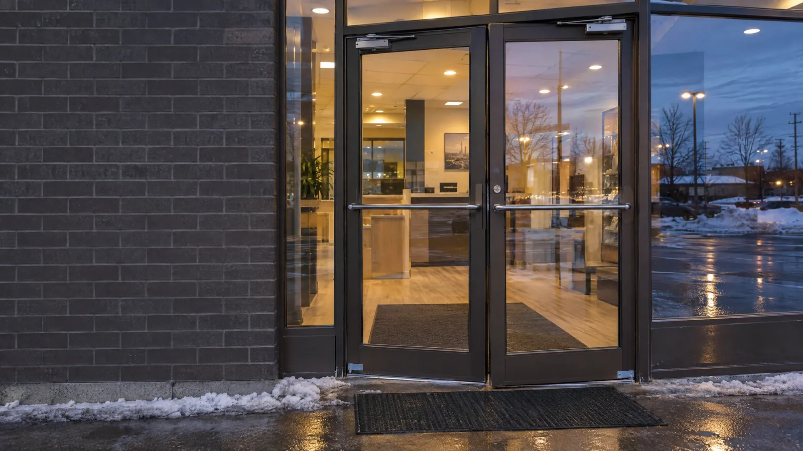 Pair of dark bronze aluminum glass entrance doors with overhead closers and push bars at a plaza unit on an icy winter evening