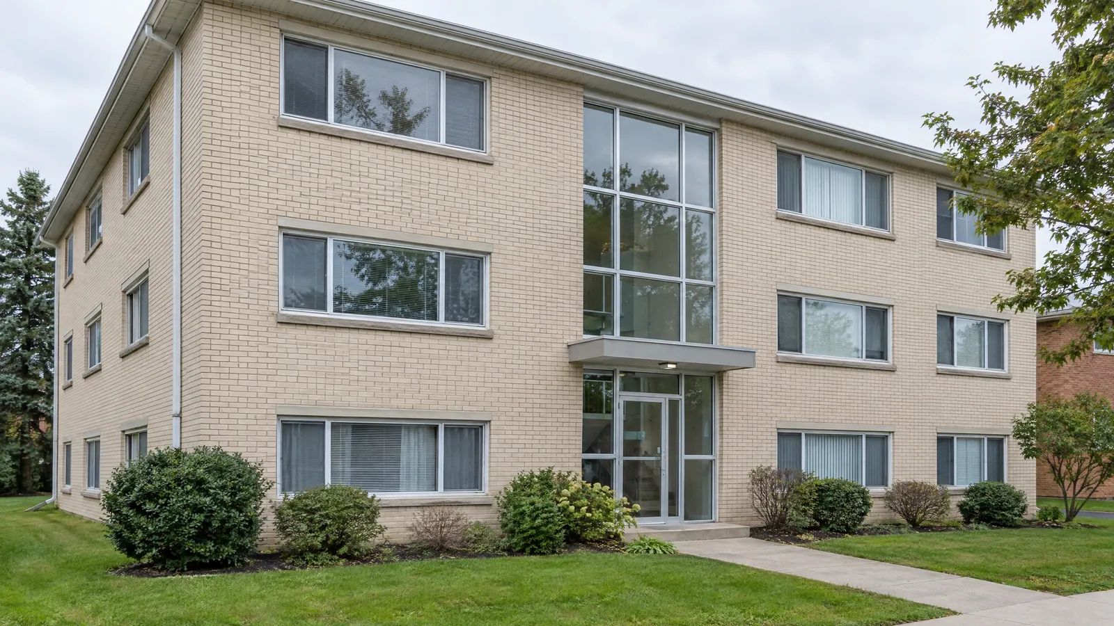 Three-storey beige brick walk-up apartment building with sliding windows on each floor and a tall glass stairwell above the front entrance