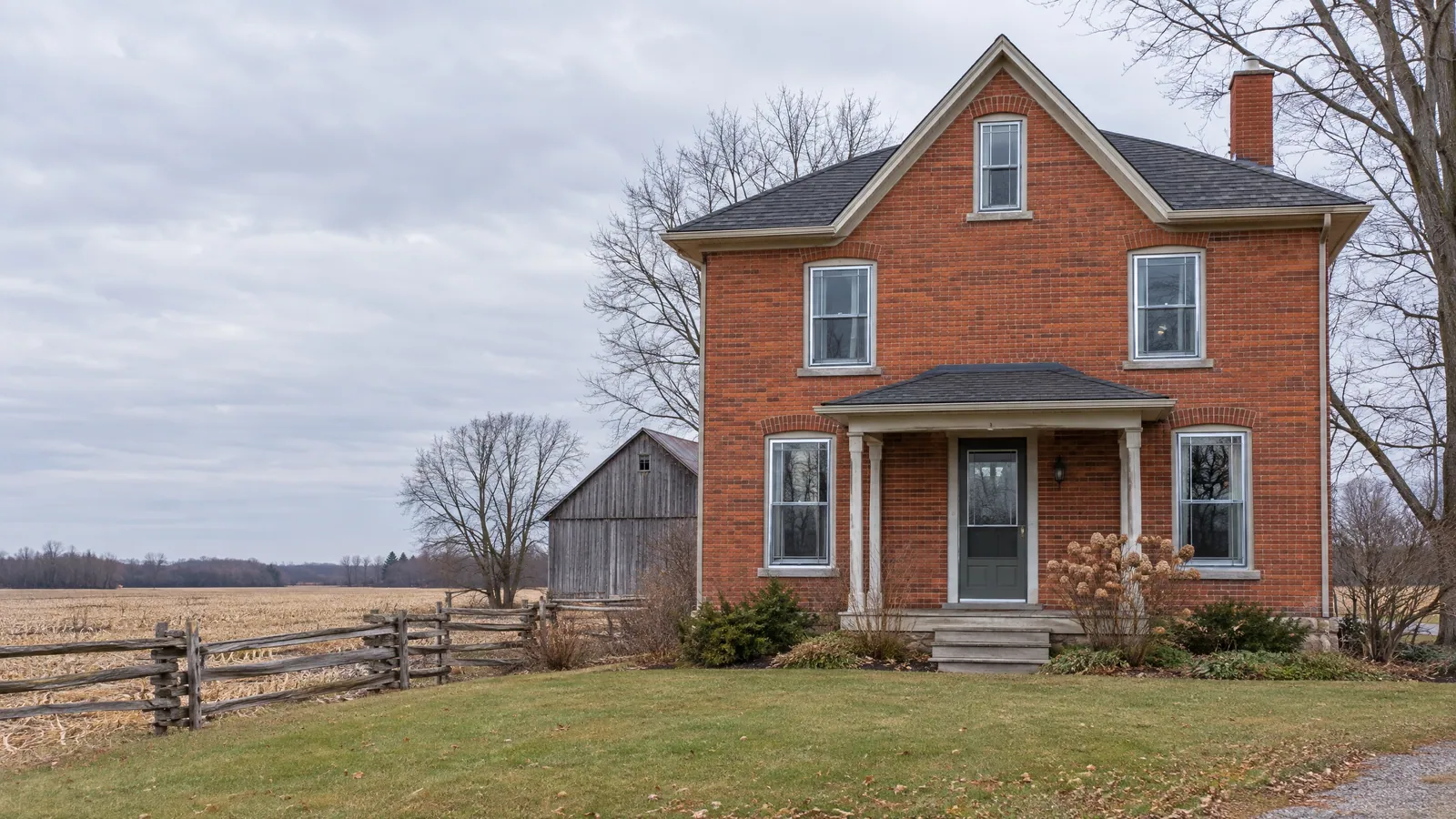 Red-brick farmhouse with storm windows over its sashes, a barn behind and harvested fields in late autumn