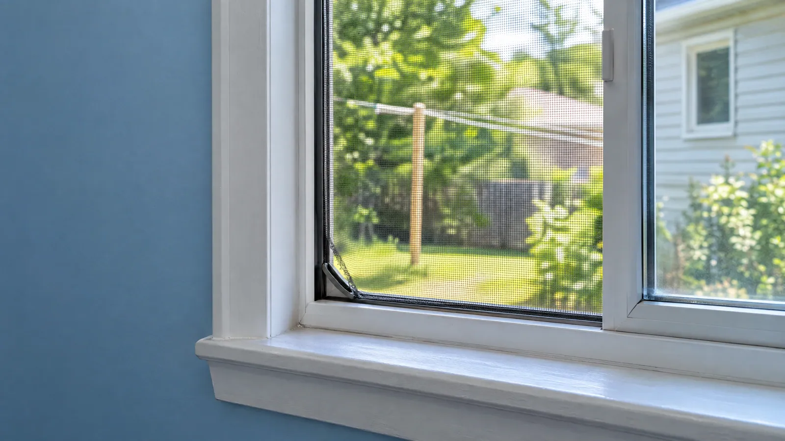 Open vinyl slider window with a corner of its charcoal insect screen torn loose, green summer trees outside